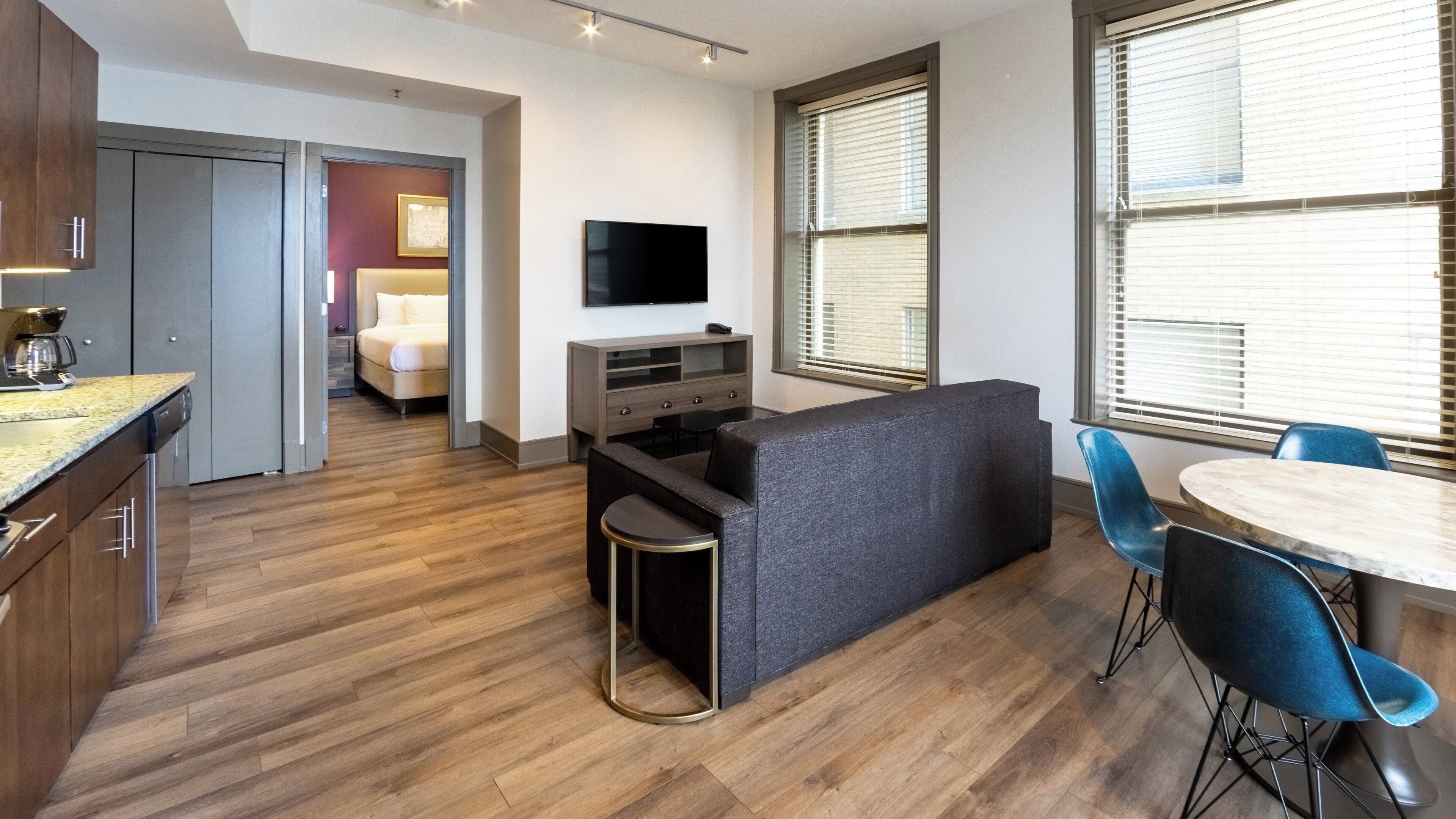 Una habitación de hotel moderna con kitchenette, un sofá, un televisor y un área de comedor con sillas azules.