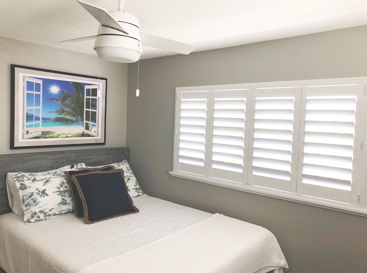 A bedroom with a bed, a ceiling fan, a framed picture of a beach scene, and white shutters on the window.