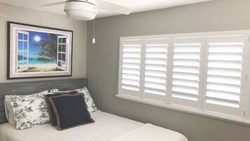 A bedroom with a bed, a ceiling fan, a framed picture of a beach scene, and white shutters on the window.