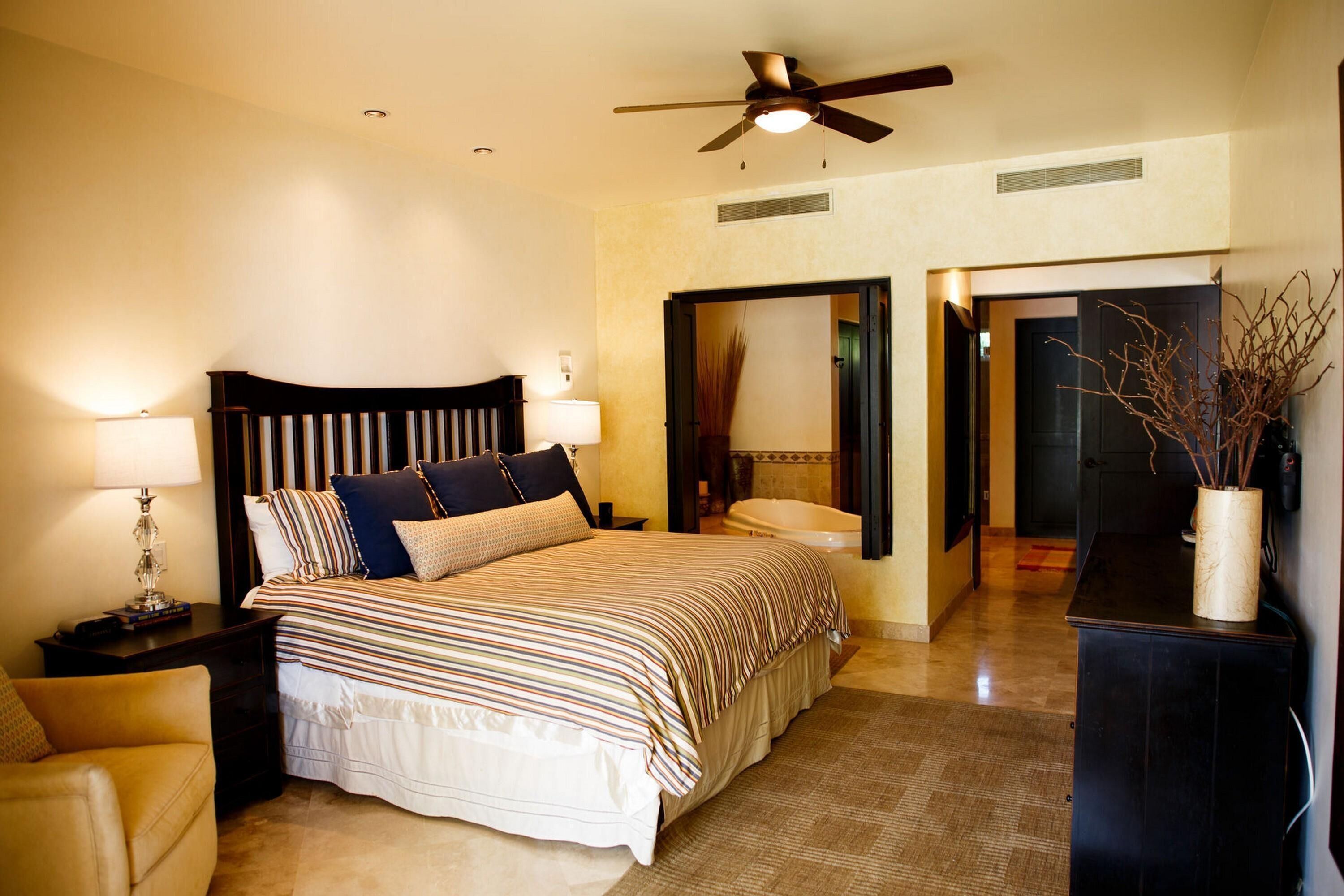 A bedroom with a bed, nightstands, a lamp, a ceiling fan, and a bathroom visible through an open door.