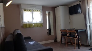 Ett litet, möblerat rum med en soffa, ett skrivbord med en stol, en tv och ett fönster med gardiner.