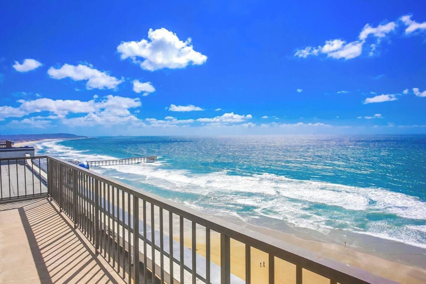 Une vue sur la côte, avec une terrasse en bois donnant sur l’océan, des vagues se brisant sur le rivage et un ciel bleu dégagé.