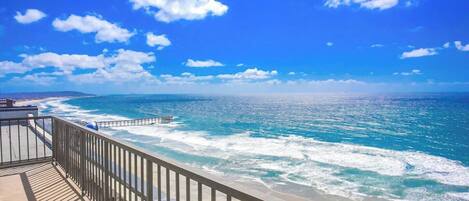 Vista costera con una terraza de madera que da al océano, olas rompiendo en la orilla y un cielo azul despejado.