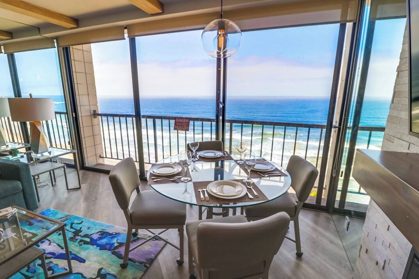 A dining area with a glass table and chairs, a view of the ocean, and a 'DANGER!' sign on the railing.