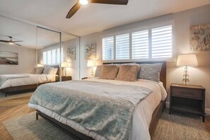 Une chambre à coucher avec un grand lit, des tables de chevet, un ventilateur de plafond et une fenêtre avec des stores.