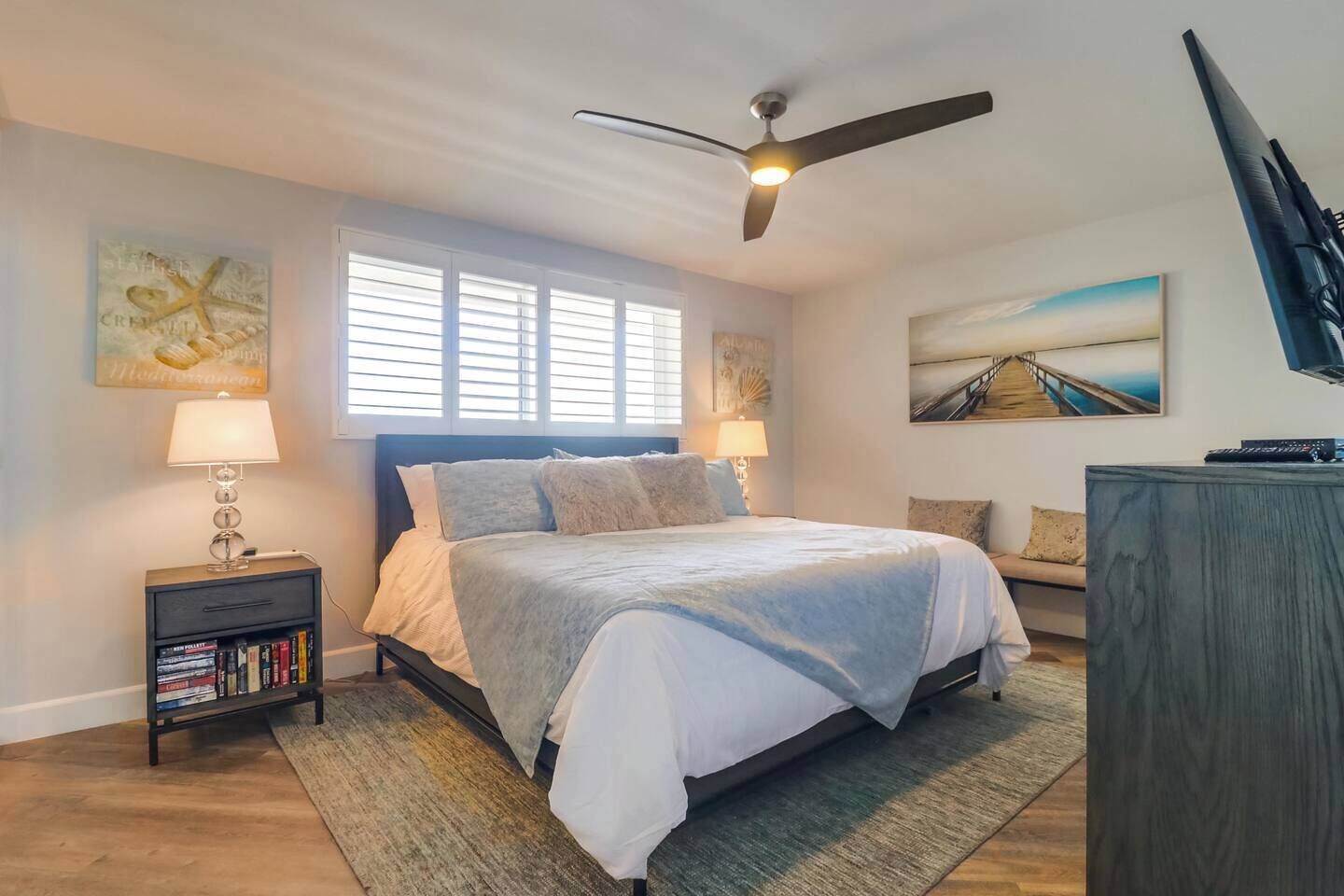 A bedroom with a bed, a nightstand, a bookshelf, a ceiling fan, and artwork on the walls.