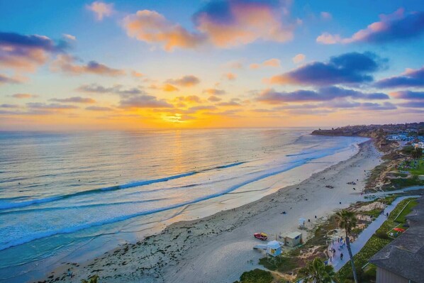 A coastal beach scene with a sunset, waves, and a path leading to a building.