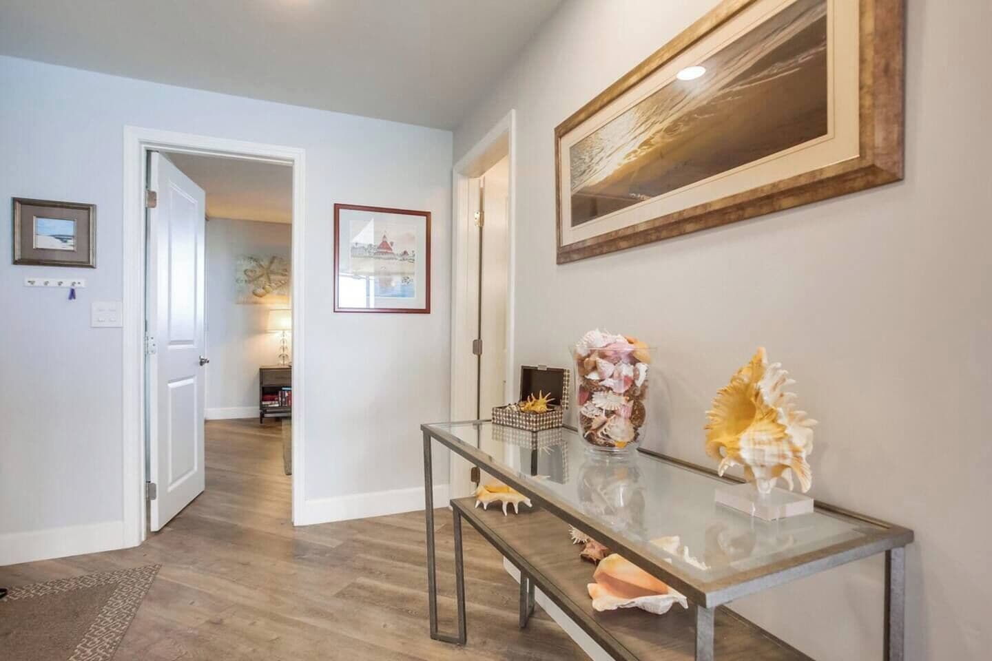 Un pasillo con una mesa consola de vidrio, obras de arte enmarcadas y una puerta que da a otra habitación.