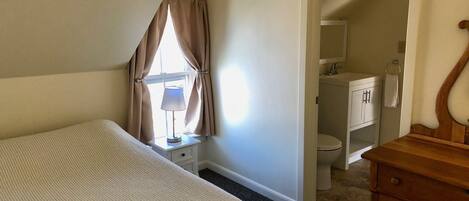 A bedroom with a bed, a bedside table with a lamp, a wooden dresser, and a bathroom visible through an open door.