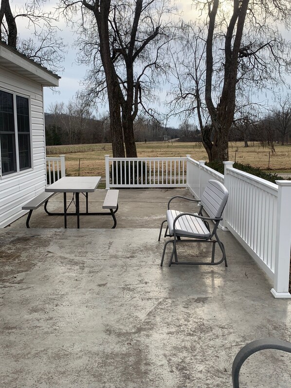 A covered patio with two metal tables and benches, white railings, and trees in the background. - Beautiful Farmhouse Stay! (Hermann)