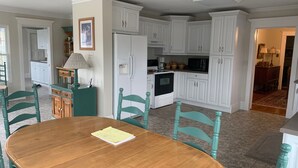 Een eetgedeelte met een houten tafel en teal gekleurde stoelen, een keuken met witte kasten en apparatuur, en een plafondventilator.