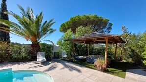 Un área de piscina con pérgola, sillones de descanso y una palmera.
