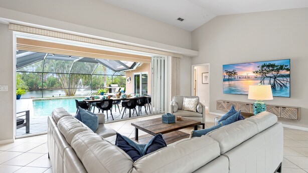 A spacious living room with a large sofa, a coffee table, and a dining area with chairs and a table. There is a large window with a view of a swimming pool and greenery outside.