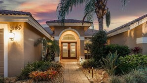 Propiedad residencial con un camino pavimentado que conduce a una puerta principal, rodeada de jardines ajardinados y palmeras.