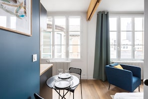 A compact room with a dining table set for two, a blue armchair, and a view of the city through large windows.