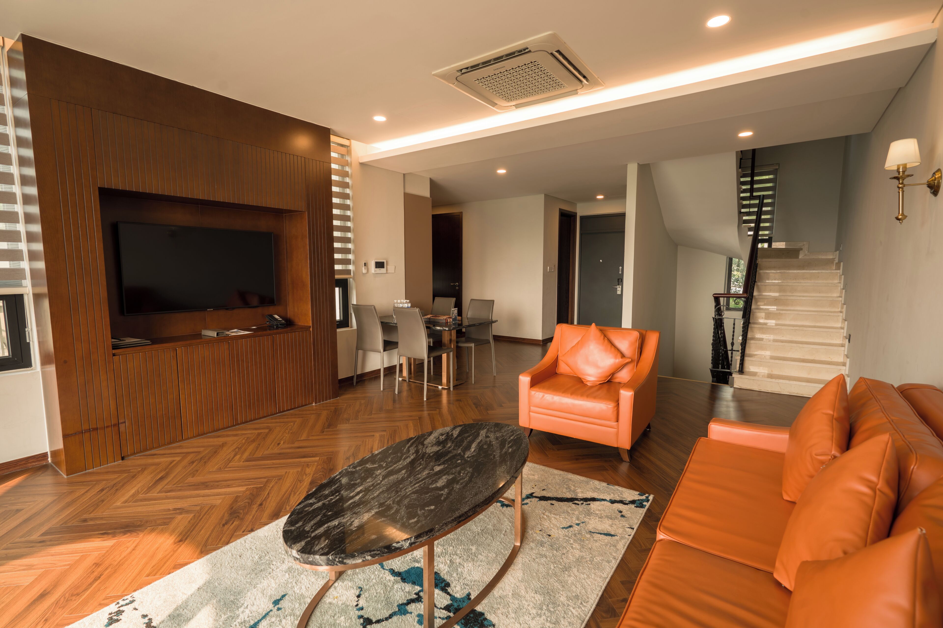 Una sala de estar moderna con un televisor de pantalla plana, una mesa de centro de mármol, un sofá de cuero naranja y una escalera que sube a otro nivel.