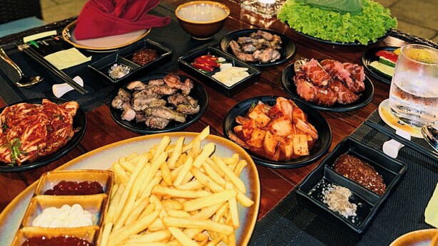 Une table à manger dressée à l’extérieur, avec divers plats, notamment de la viande, des légumes et des frites, et en arrière-plan, une piscine et des éclairages.