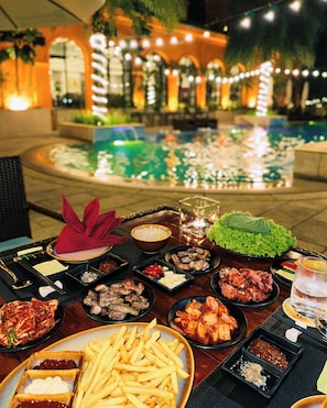 A dining table with various dishes, including meat, vegetables, and French fries, set outdoors with a pool and lights in the background. - Wyndham Sky Lake Resort and Villas (Hanoi)