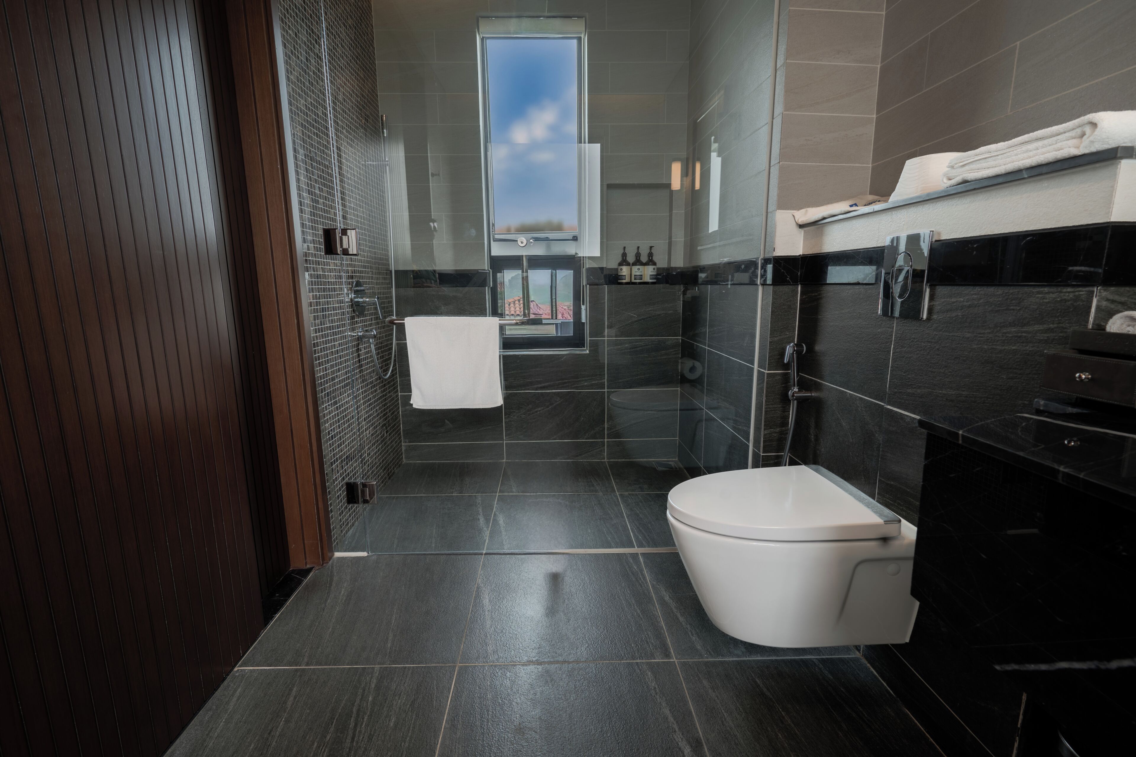 A modern bathroom with a toilet, sink, and shower area, featuring dark tiles and a visible window.
