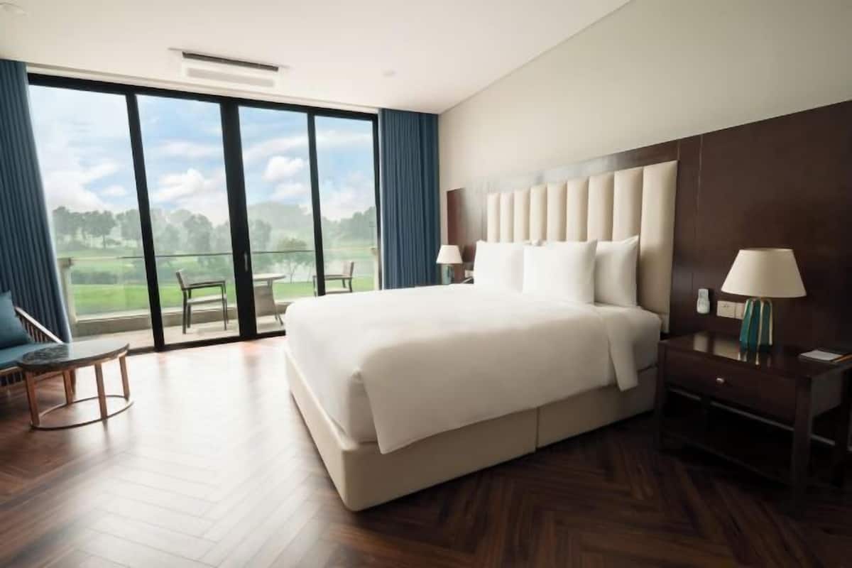 A hotel room with a large bed, a desk, a chair, and a view of a golf course through large windows.