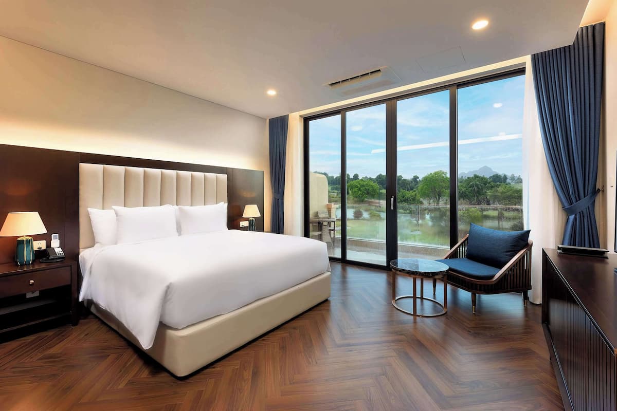 A modern hotel room with a large bed, a seating area with a chair, a small table, and a large window offering a view of the outdoors.