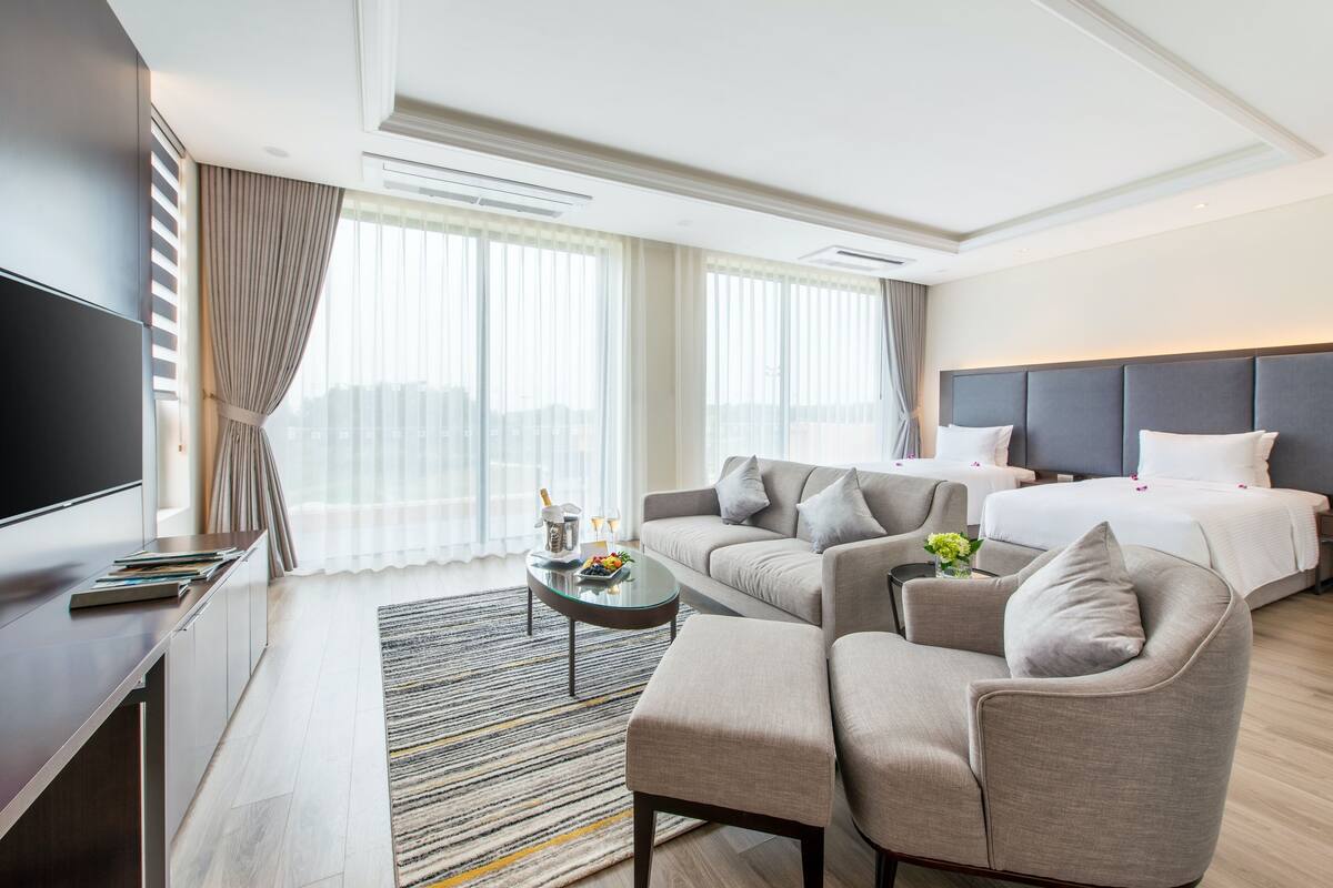 A hotel room with a bed, sofa, armchair, TV, and a small table with a vase of flowers.