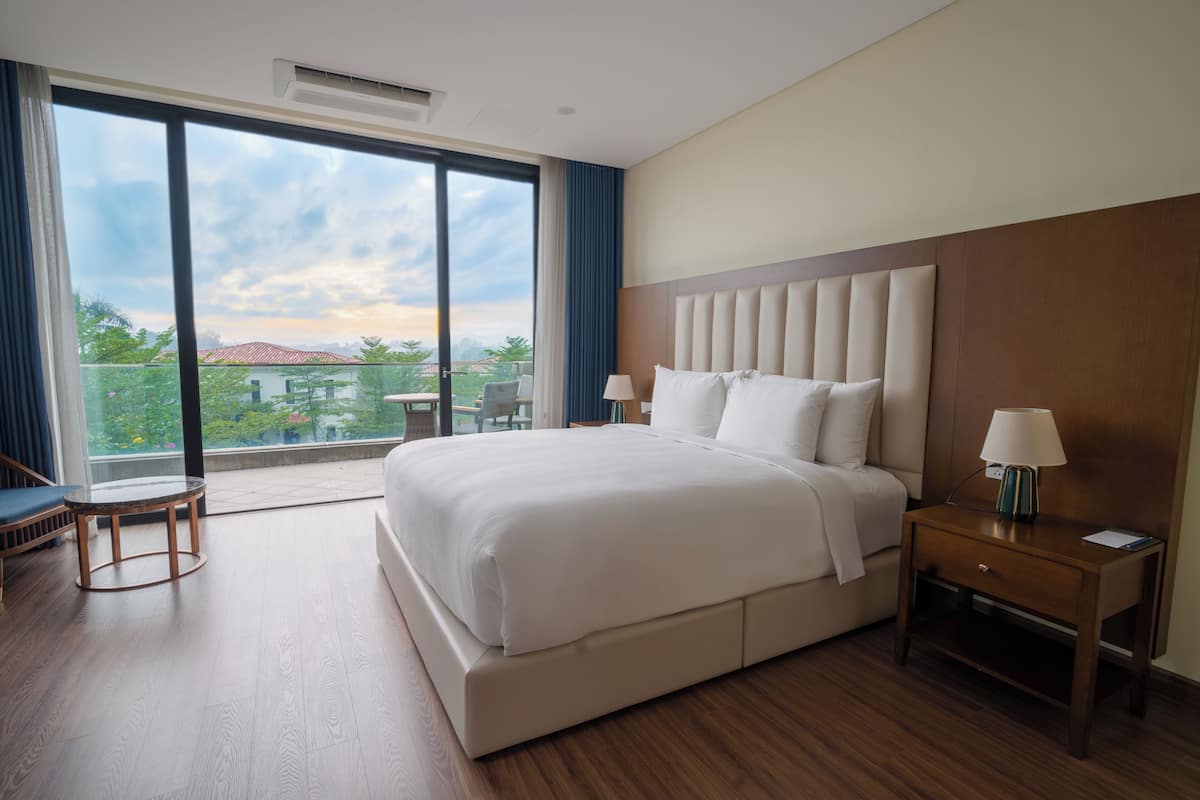 A hotel room with a large bed, a bedside table with a lamp, a small round table, and a large window offering a view of greenery and buildings.