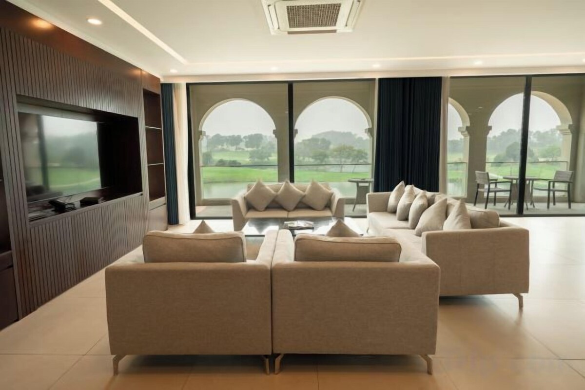 A modern living room with a large flat-screen TV, a beige sofa set, and a view of a green landscape through large windows.