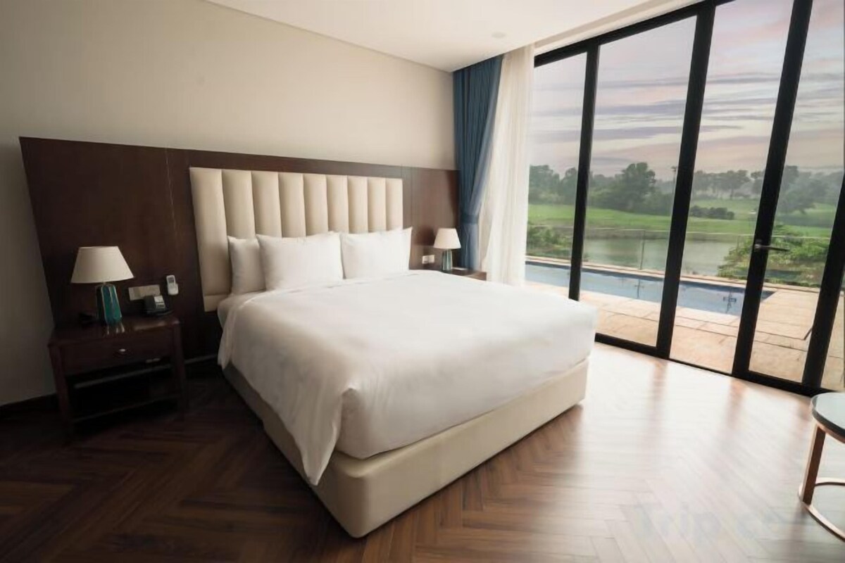 Una cama grande con cabecera acolchada, una mesita de noche con lámpara, y vista a un cuerpo de agua a través de amplias ventanas.