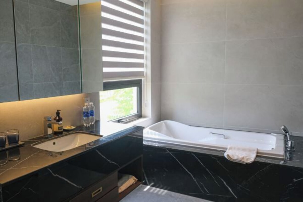 A modern bathroom with a black marble vanity, a white bathtub, and a window with blinds.