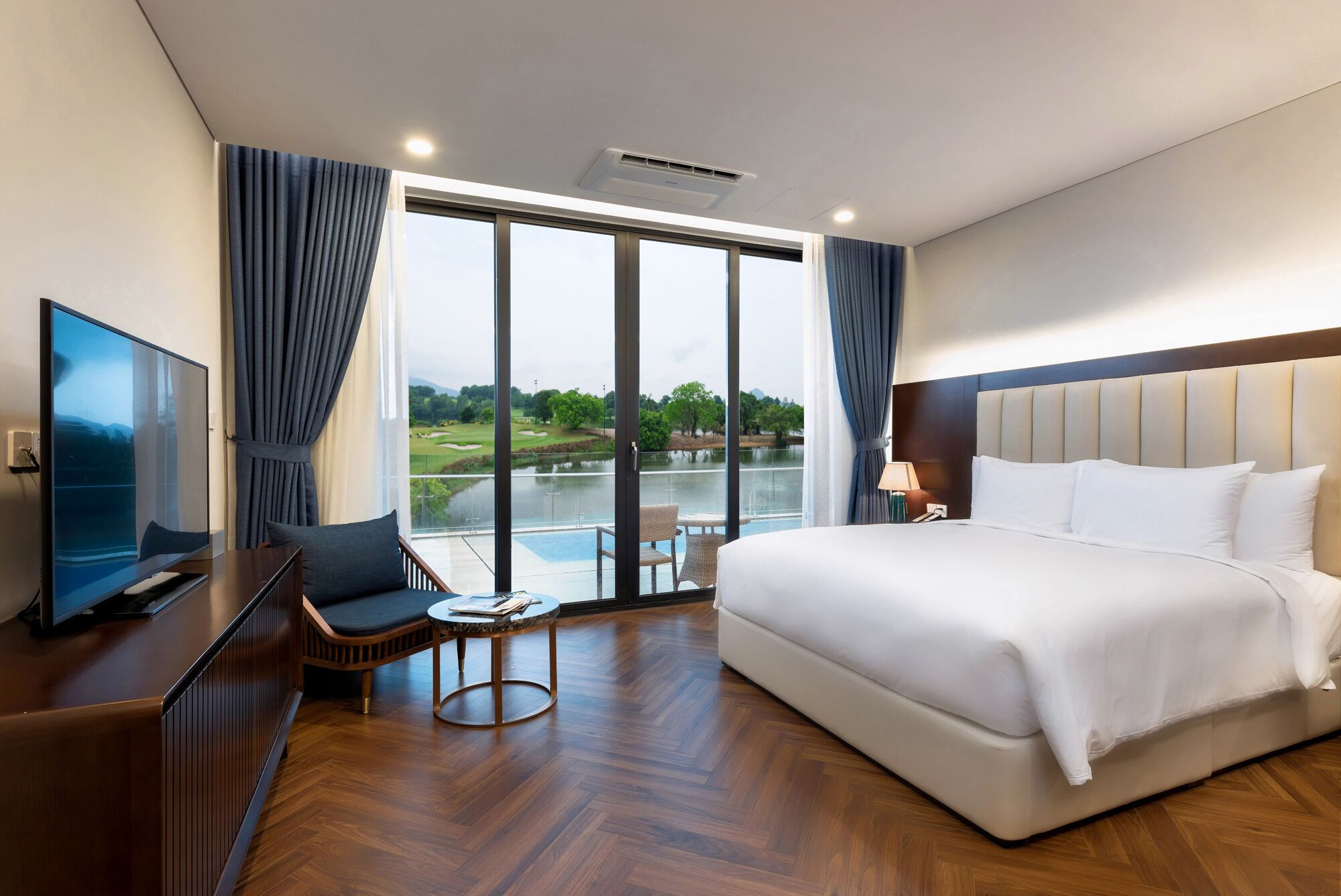A hotel room with a large bed, a television, a small table, and a view of a golf course and lake.