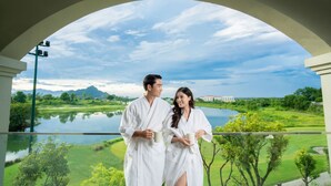 Un couple, vêtu de longues robes blanches, se tient sur un balcon d’où l’on aperçoit un lac, un terrain de golf et des montagnes.