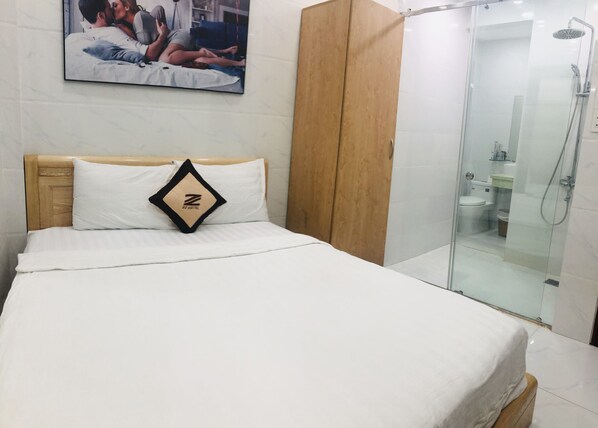 A hotel room with a bed, a wooden wardrobe, a glass shower enclosure, and a framed picture on the wall.