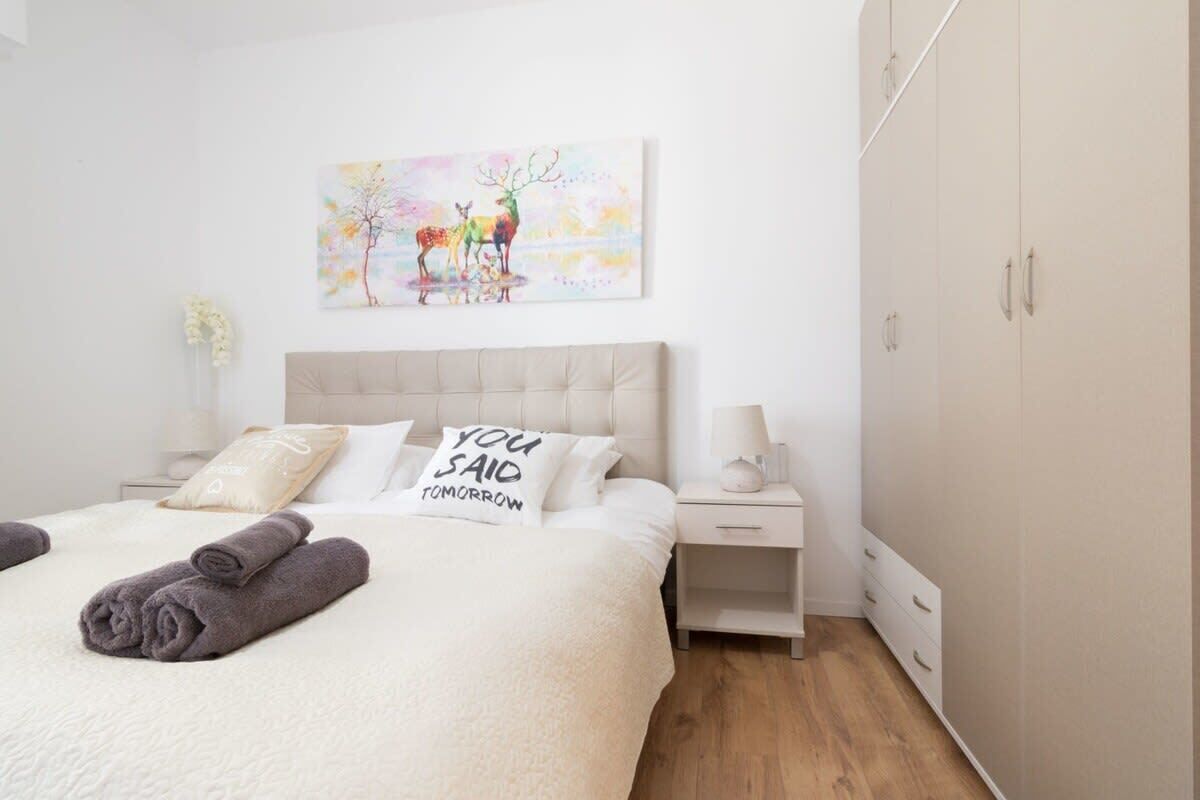A neatly made bed with a pillow that reads "YOU SAID TOMORROW," a bedside table with a lamp, and a wardrobe.