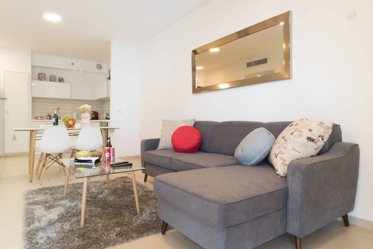 A modern living room with a grey sofa, a glass coffee table, and a small rug. There is a kitchen in the background with white cabinets and a dining area with white chairs.