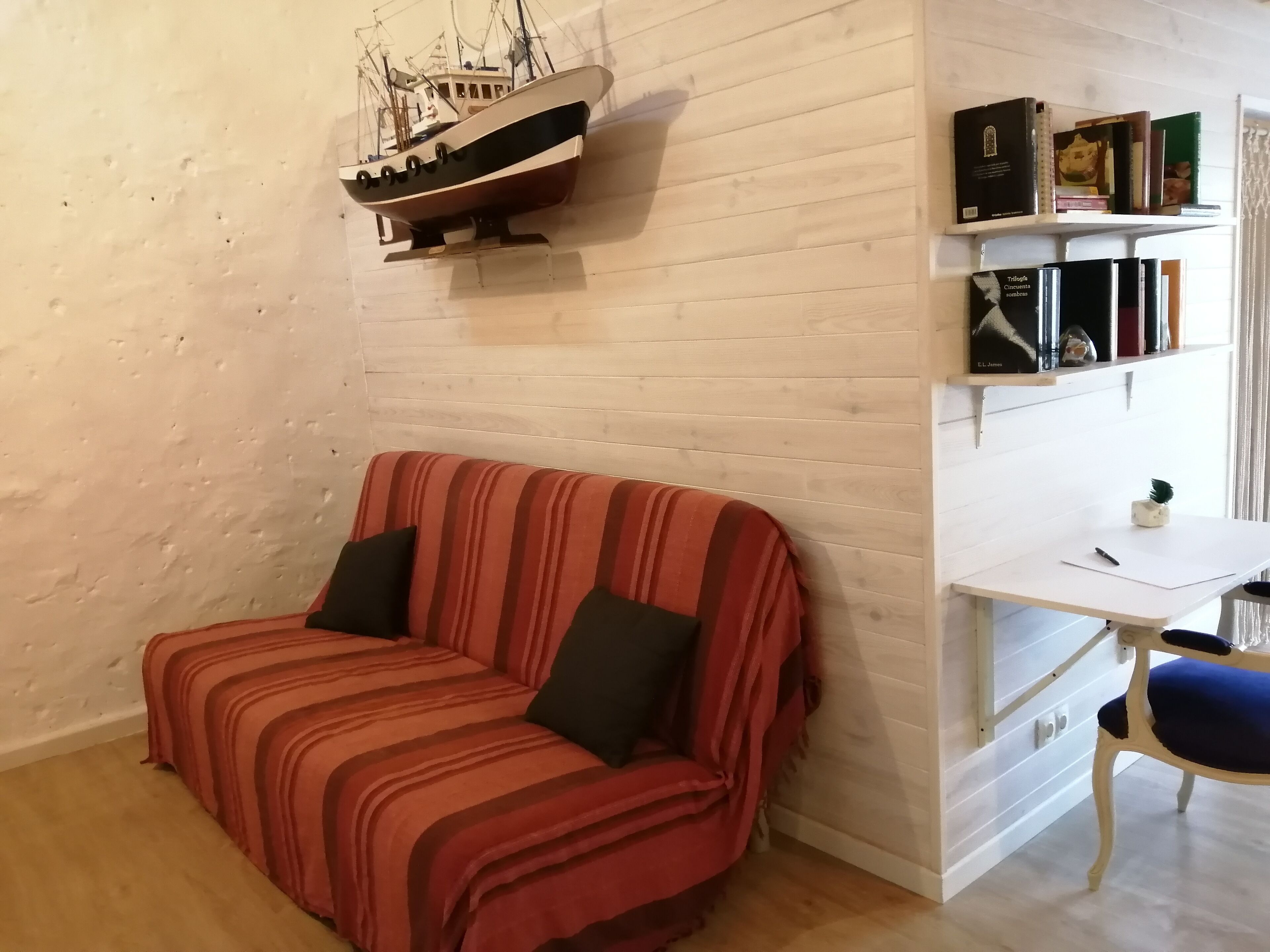 Ein Zimmer mit einem gestreiften Sofa, einem Schreibtisch mit Stuhl, einem Bücherregal und einem Modellschiff an der Wand.