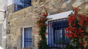A stone building with a window adorned with red flowers and blue bars. There is a table and two wicker chairs outside. - Comfortable Fisherman's House (Llançà)
