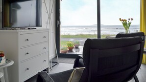 A modern room with a television on a white cabinet, a large window with a view of the sea, and a chair facing the window. - The Isle View Nest - UK13547 (Lower Harrapool, near Broadford)