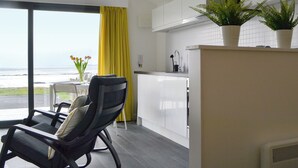 A modern kitchen with white cabinets, a dining area with a black chair, a yellow curtain, and a view of the ocean. - The Isle View Nest - UK13547 (Lower Harrapool, near Broadford)