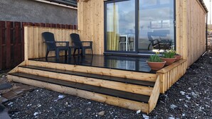 A wooden cabin with a glass door, a small outdoor seating area with chairs, and a gravel ground. - The Isle View Nest - UK13547 (Lower Harrapool, near Broadford)