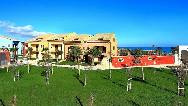 A large, multi-story hotel with a Mediterranean architectural style, featuring a central courtyard with palm trees and a view of the ocean. - Open space apartment (Licata)