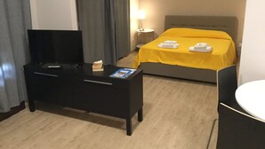 A hotel room with a bed, a TV on a stand, a bedside table with a lamp, and a small table with a magazine. - Open space apartment (Licata)
