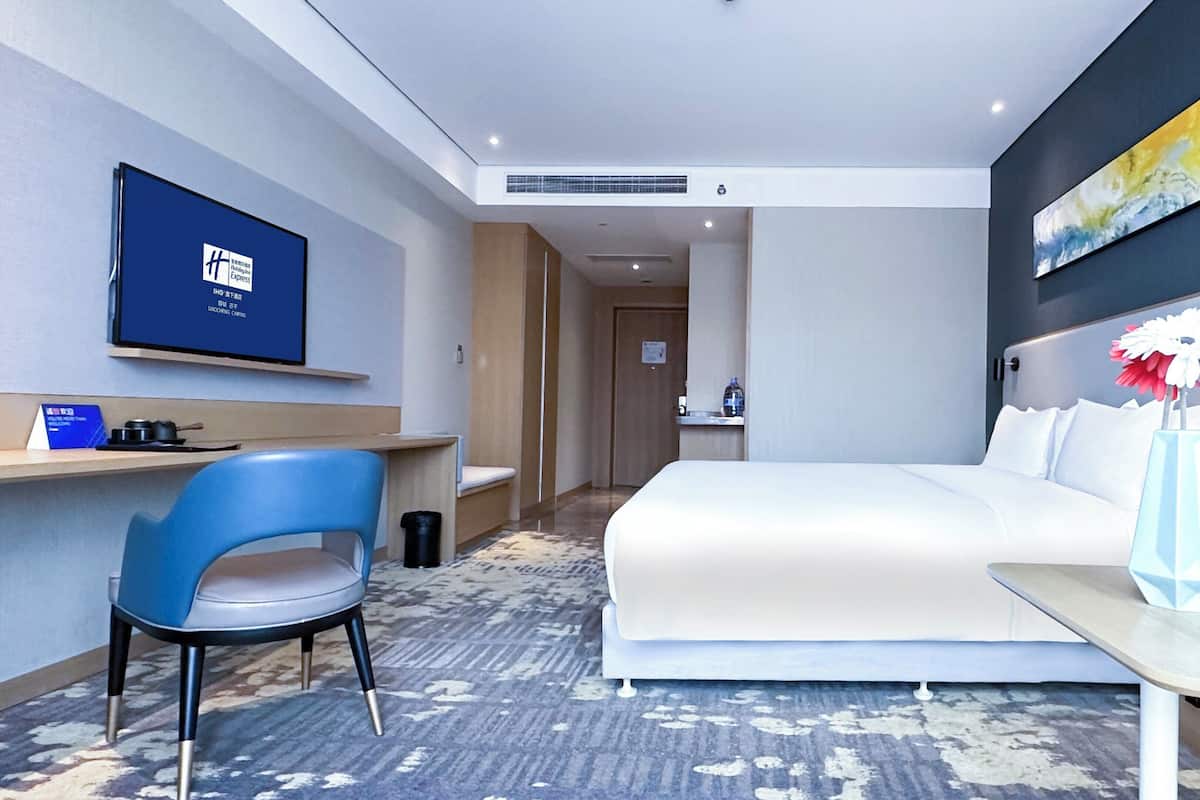 A hotel room with a bed, a desk, a chair, a TV, a vase with flowers, and a painting on the wall.
