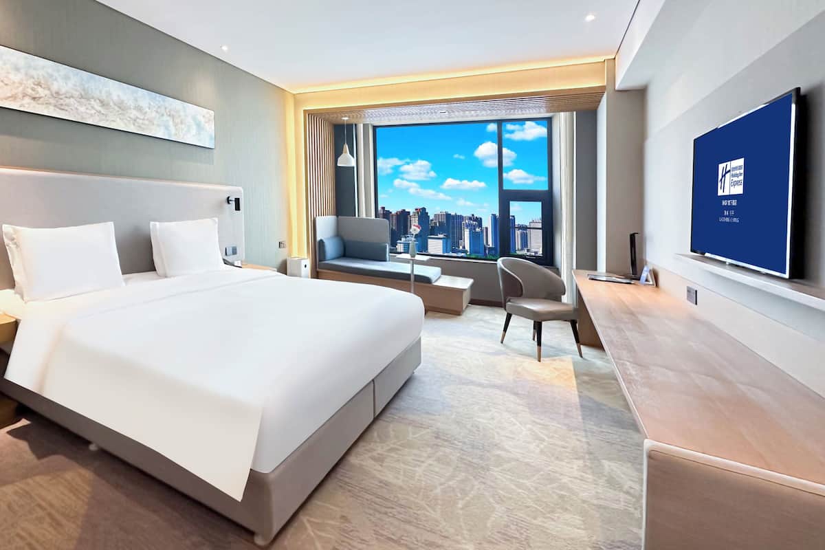 A modern hotel room with a large bed, a seating area with a view of the city, and a flat-screen TV on the wall.