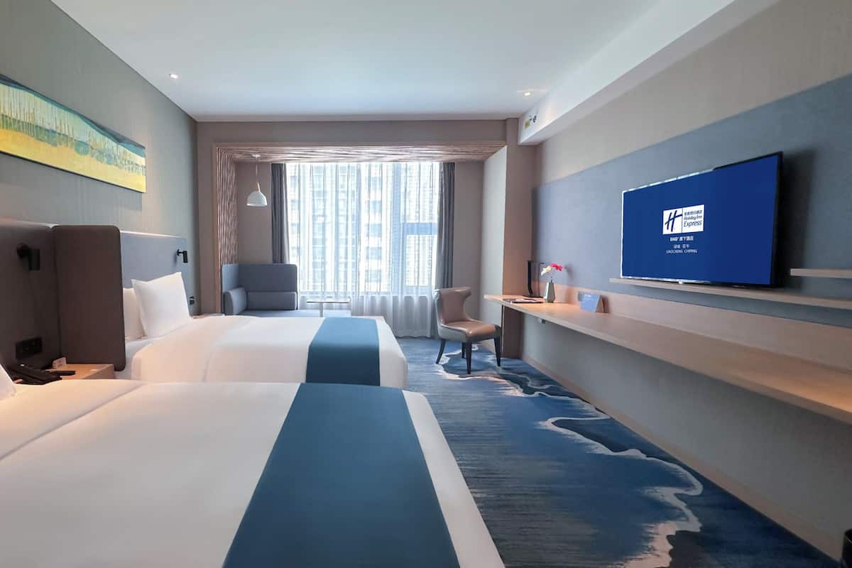 A hotel room with two beds, a TV, a desk, and a large window with curtains.