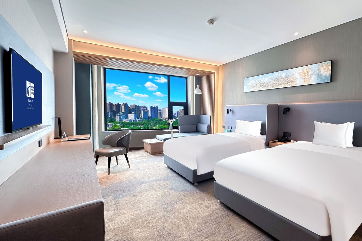 A hotel room with two beds, a desk, a chair, and a large window with a city view.