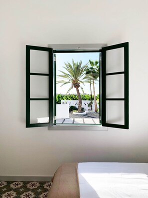 A room with a bed, a window with open shutters, and a view of palm trees and a wall with greenery.