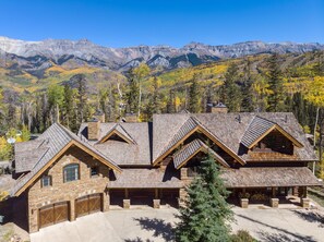 A log cabin with a stone foundation, multiple chimneys, and a large garage. It is surrounded by a mountainous landscape with autumn foliage. - Hilltop Hideaway - Expansive Views From This Private Estate, Hot Tub, Pool Table (Telluride)