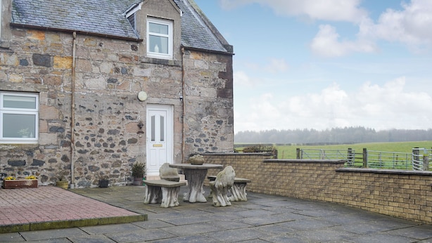 A stone house with a tiled roof, a white door, and a small window. There is a paved patio with a table and chairs. - 4 bedroom accommodation in Fenwick, near Kilmarnock (Fenwick, near Kilmarnock)