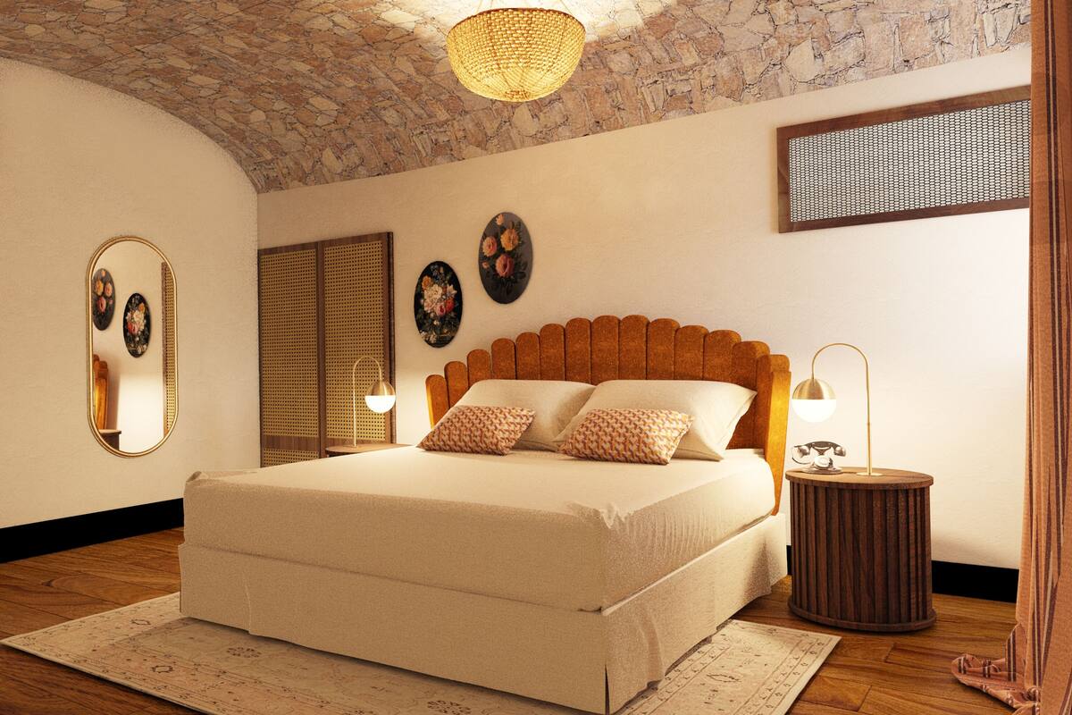 A bedroom with a stone ceiling, a bed with patterned pillows, a round side table with a lamp, and a mirror on the wall.